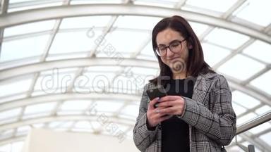 一个年轻的女人拿着电话站着。 一个女孩用智能手机对着一个玻璃屋顶。 一个忙碌的人正在发短信